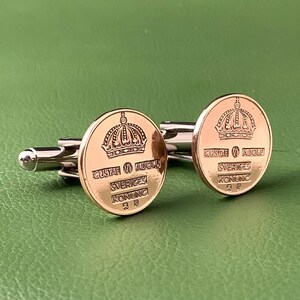 May include: A pair of gold-toned cufflinks with a circular design featuring a crown and text. The cufflinks have a silver-colored backing and are displayed on a green surface. The text on the cufflinks reads "GUSTAF VI ADOLF, SVERIGES KONUNG".
