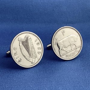 May include: Silver cufflinks featuring Irish coins. One cufflink displays a harp and the year 1959, while the other shows a bull and the inscription "1s scilling". The cufflinks are set against a blue background.