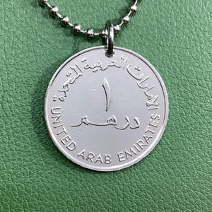 May include: Silver necklace with a round pendant featuring the text "United Arab Emirates" in English and Arabic. The pendant also has the number "1" on it.