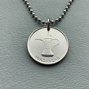 May include: A silver necklace with a silver coin pendant featuring a traditional Arabic coffee pot design. The coin has the inscription "1310-1990" on it.