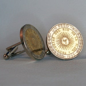 May include: A pair of cufflinks featuring a gold-colored coin with the inscription "UN PESO DEL RIO DE LA PLATA PROVINCIA 1995". The coin has a sun with a face in the center.
