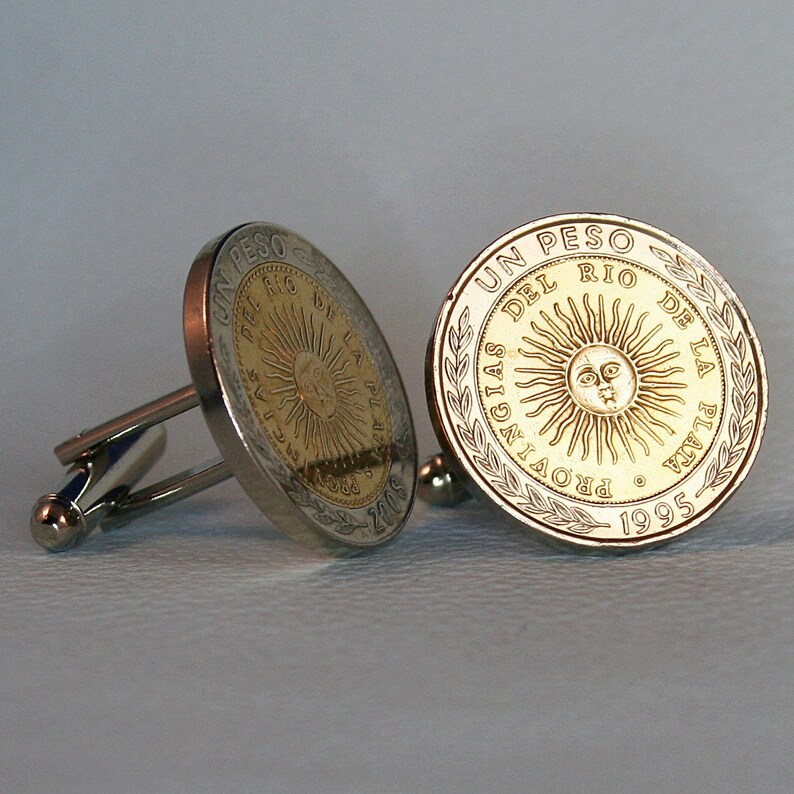 May include: A pair of cufflinks featuring a gold-colored coin with the inscription "UN PESO DEL RIO DE LA PLATA PROVINCIA 1995". The coin has a sun with a face in the center.