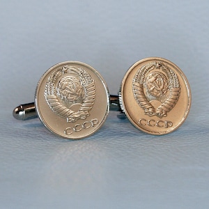 May include: A pair of silver cufflinks featuring a circular design with a hammer and sickle emblem surrounded by a wreath and the inscription "CCCP".