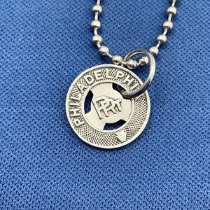May include: A silver-colored pendant necklace with a round charm. The charm has the word "PHILADELPHIA" around the edge and a logo in the center. The necklace chain is made of small, round beads.