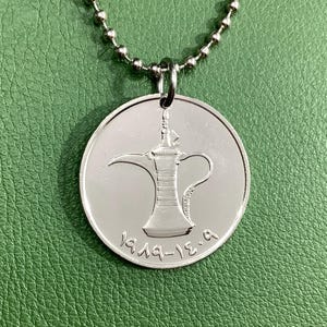 May include: Silver necklace with a round pendant featuring a detailed engraving of a traditional Arabic coffee pot. The pendant has the inscription "1919-13.9" on the bottom.