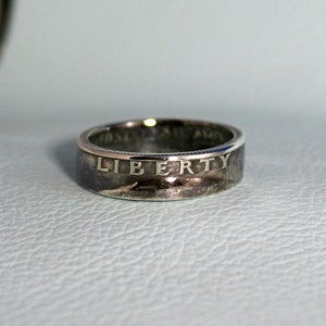 May include: A silver ring made from a US quarter coin. The ring features the inscription "LIBERTY" on the band.