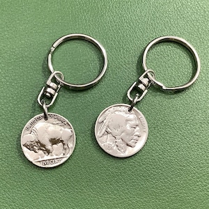 May include: Two silver keychains, each featuring a vintage buffalo nickel coin. One coin shows a buffalo, the other a Native American profile. The keychains have silver rings and clasps, set against a green background.
