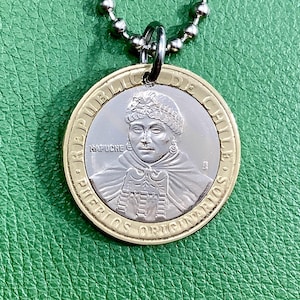 May include: A gold and silver pendant necklace featuring a portrait of a person with a headdress. The pendant has the text "REPUBLICA DE CHILE" and "PUEBLOS ORIGINARIOS" around the edge.