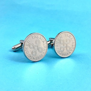 May include: A pair of silver cufflinks with a circular design. Each cufflink features a coin with the text "SUOMEN TASAVALTA" and the year 1955 and 1961. The cufflinks are set against a light blue background.