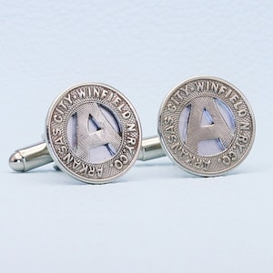 Könnte beinhalten: Ein Paar silberne Manschettenknöpfe mit einem runden Design mit dem Text "Arkansas City Winfield Ry Co" und dem Buchstaben "A" in der Mitte.
