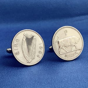 May include: Silver cufflinks featuring Irish coins. One cufflink displays a harp and the year 1954, while the other shows a bull and the inscription "1s". The cufflinks are set against a blue background.