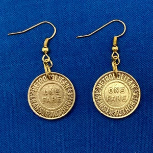 May include: A pair of gold-colored earrings. Each earring features a circular charm with the words "ONE FARE" and "METRO TRANSIT AUTHORITY" engraved on it. The earrings have a simple hook design and hang against a blue background.