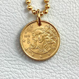 May include: A gold-colored pendant necklace featuring a coin with a woman's profile, stars, and the year 2010. The necklace has a ball chain. The coin pendant is approximately 1 inch in diameter.
