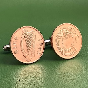 May include: Copper cufflinks featuring Irish coins. One cufflink displays a harp and the word "Eire" with the year 1995. The other cufflink shows a bird-like design and the number "1P". The cufflinks have a silver-colored metal backing.