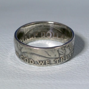May include: A silver ring made from a 1963 half dollar coin. The ring features the inscription "IN GOD WE TRUST" and "HALF DOLLAR" on the band.
