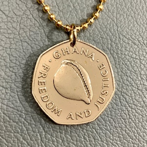 May include: Gold necklace with a round pendant featuring the words "Ghana Freedom and Justice" and a stylized image of a shell.