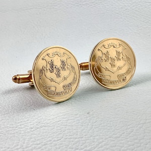 May include: Gold-toned cufflinks with a circular design featuring a coat of arms and the Latin phrase "QUI TRANSTULIT SUSTINET." The cufflinks have a classic, elegant appearance.