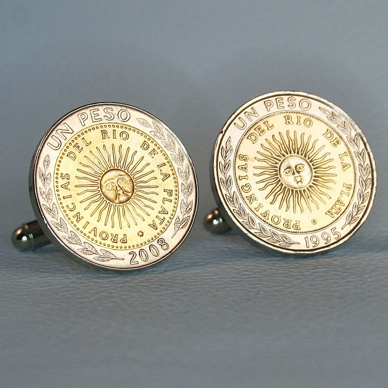 May include: A pair of silver cufflinks featuring a gold coin design. Each coin features a sun with a face and the text "UN PESO DEL RIO DE LA PLATA" around the edge. One coin has the year "2008" and the other has the year "1995".