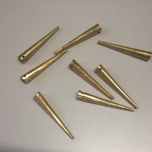 May include: A group of eight gold-colored metal tacks with pointed tips. The tacks are scattered on a light gray surface.