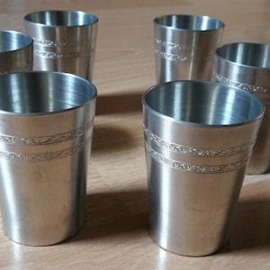 May include: Six brushed metal shot glasses. Each glass has a decorative band near the top and is set on a wooden surface. The glasses are cylindrical with a slightly flared top.