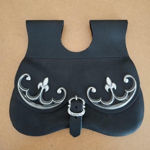 Könnte beinhalten: Schwarze Ledertasche mit silberfarbenen Zierelementen. Die Tasche hat einen Schnallenverschluss und ein silberfarbenes Dekor. Die Oberseite hat zwei Schlaufen zur Befestigung. Die Tasche ist wahrscheinlich zum Tragen kleiner Gegenstände gedacht.