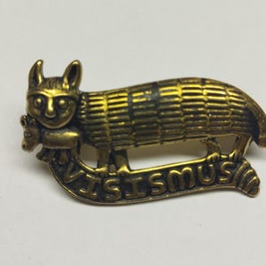 Peut inclure: Épingle en laiton de style vintage représentant un chat stylisé tenant un petit animal. Le corps du chat est texturé avec des lignes horizontales. L'épingle comprend le mot "VISISMUS" dans une bannière incurvée.