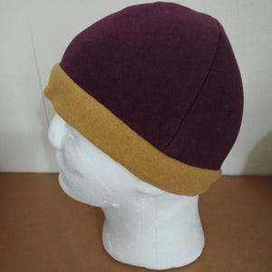 May include: A burgundy beanie with a mustard yellow brim is displayed on a white mannequin head. The hat is made of a soft, textured material. The brim is a contrasting color, adding a stylish accent to the hat.