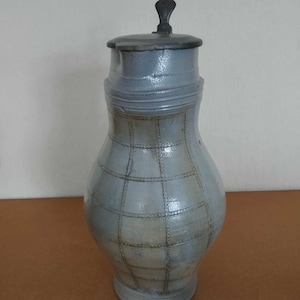 May include: A grey ceramic jug with a pewter lid and a chequered pattern. The jug has a rounded body and a narrow neck.