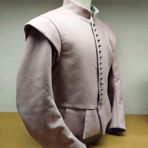 May include: A pale mauve doublet with a high collar and shoulder pads. The garment has long sleeves and small, silver buttons down the front. The bottom of the doublet features a peplum design. The garment is displayed on a mannequin.