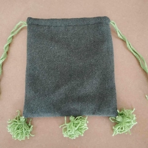 Könnte beinhalten: Eine dunkelgraue Kordeltasche mit grünen Quasten. Die Tasche ist aus einem weichen, flauschigen Material.