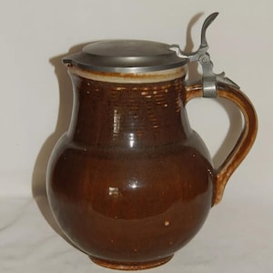 May include: A brown ceramic beer stein with a metal hinged lid and handle. The stein has a rounded body and a textured surface near the top. The handle and lid are silver-coloured.