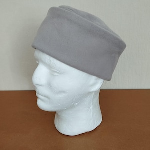 Puede incluir: Un sombrero gris claro con una corona rectangular y un ala ligeramente inclinada. El sombrero se exhibe en una cabeza de maniquí blanca, mostrando su forma y diseño. El sombrero está hecho de un material suave.