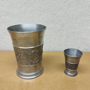 May include: Two pewter drinking vessels. The larger one has a flared top and a decorative band with a floral design. The smaller cup has a similar design. Both are made of a silver-coloured metal and sit on a light brown surface.