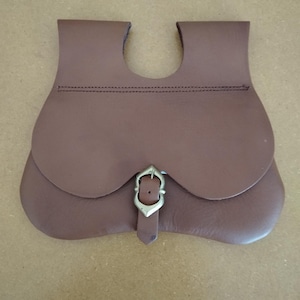 Könnte beinhalten: Eine braune Ledertasche mit gebogenem Deckel und Messing-Schnallenverschluss. Die Tasche hat eine einzigartige Form mit einem Ausschnitt oben und ist mit dunklem Faden genäht. Das Design ist schlicht und funktional.