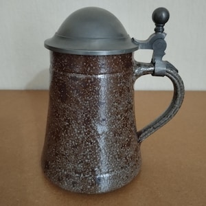 May include: A brown ceramic beer stein with a pewter lid and handle. The stein has a speckled brown glaze.