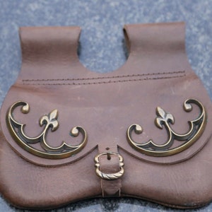 Könnte beinhalten: Braune Ledertasche mit dekorativen Messingakzenten. Die Tasche hat einen Schnallenverschluss und zwei Schlaufen oben. Die Messingakzente weisen ein Fleur-de-Lis-Design auf.