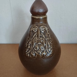 May include: A brown ceramic bottle with a conical stopper. The bottle features a detailed relief of a bearded face. The bottle is approximately 20 cm tall.