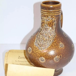 Puede incluir: Jarra de cerámica marrón con asa, con un relieve detallado de una cara barbuda y motivos florales decorativos. La jarra tiene un cuerpo redondeado y un cuello estrecho. Un certificado es parcialmente visible.