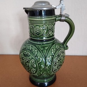 Può includere: Un boccale da birra in ceramica verde con un motivo floreale decorativo e un coperchio incernierato in metallo. Il manico e il corpo sono verdi, con accenti neri sulla base e sul bordo. Il boccale ha una forma bulbosa e un design in rilievo dettagliato.