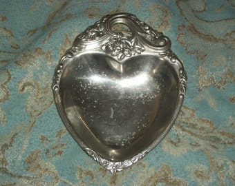 May include: Silver-toned heart-shaped dish with ornate floral and scroll detailing. The dish has a handle at the top and a slightly concave interior. The edges are decorated with small flowers and leaves. A decorative piece for serving or display.