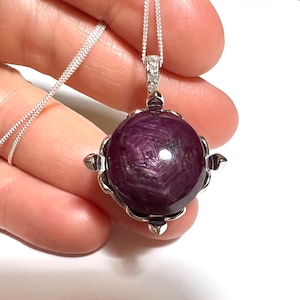May include: A silver pendant necklace featuring a large, round, dark red ruby gemstone set in a silver bezel. The ruby has a star-like pattern and is surrounded by small silver accents.