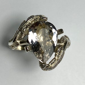 Może przedstawiać: Srebrny pierścionek z przezroczystym kamieniem szlachetnym w kształcie gruszki osadzonym w wzorze piór.