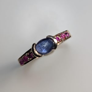 Puede incluir: Un anillo de plata con una gran gema azul ovalada en el centro, flanqueada por gemas rosas más pequeñas. El anillo tiene un diseño elegante y moderno con un acabado pulido, que muestra los colores vibrantes de las piedras.