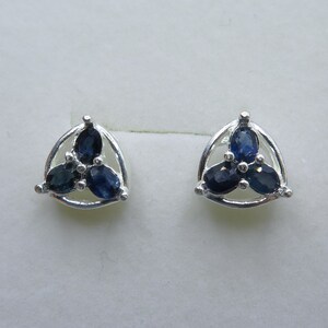 May include: A pair of silver stud earrings. Each earring has a triangular frame holding three oval, dark blue gemstones. The gemstones are clustered, forming a floral or geometric shape. The earrings are displayed on a white surface.
