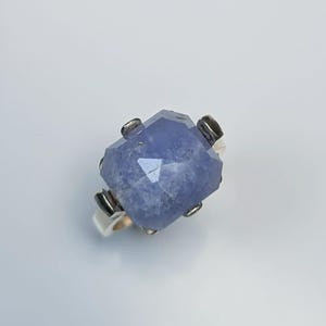 Puede incluir: Anillo con una gema azul claro y una banda de plata. La gema rectangular tiene múltiples facetas y está sujeta por cuatro garras de plata. El anillo está sobre un fondo blanco. Joya elegante y atemporal.