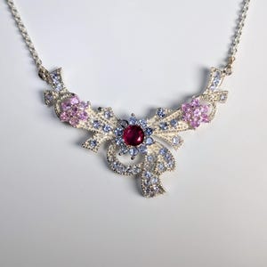 May include: A silver necklace with a bow-shaped pendant. The pendant features a large, round, red gemstone at the center, surrounded by smaller blue and pink gemstones. The necklace has a delicate chain.