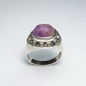 Puede incluir: Un anillo de plata con una gran piedra preciosa púrpura ovalada en el centro. El anillo está adornado con piedras preciosas más pequeñas, de color verde oscuro y azul, alrededor de la piedra principal, engastadas en una banda de plata detallada. El anillo tiene un acabado pulido.
