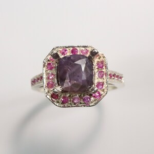 Puede incluir: Un anillo de plata con una gran piedra preciosa cuadrada de color púrpura rodeada por un halo de piedras preciosas rosas más pequeñas.