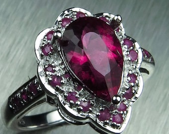 May include: A pear-shaped ruby gemstone ring set in silver with a floral design and a halo of smaller rubies.