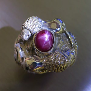 May include: A silver ring featuring a large, oval, star ruby gemstone. The ring's setting is intricately sculpted with a detailed, organic design. The ruby is a deep reddish-purple color, with a prominent six-ray star.
